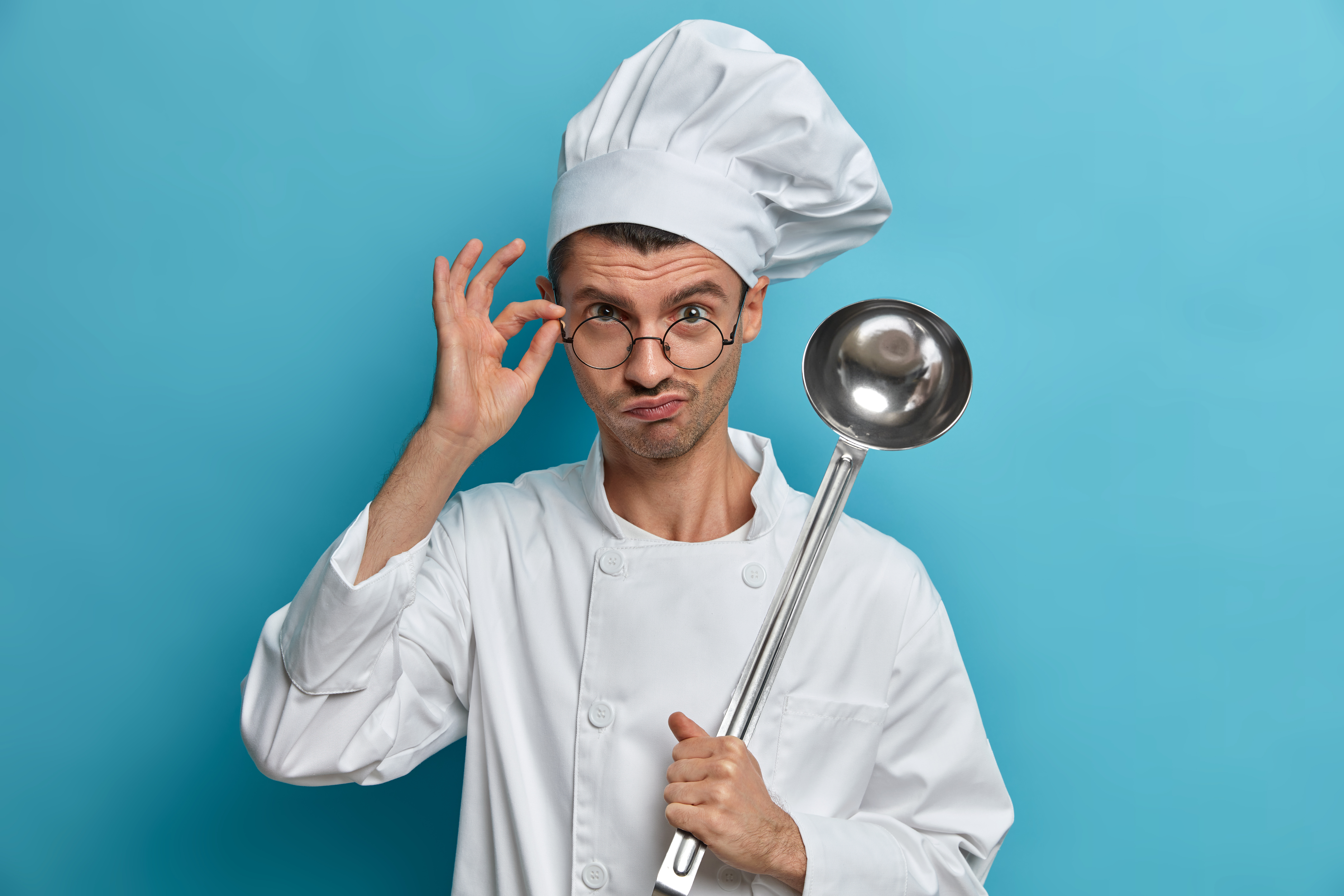 chef cook poses commercial kitchen looks seriously through glasses holds ladle prepares meal ready cook dish listens advice