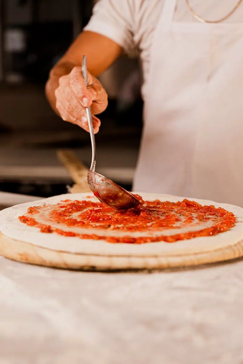 Chef spreading tomato sauce on pizza dough in a kitchen setting with a spoon.